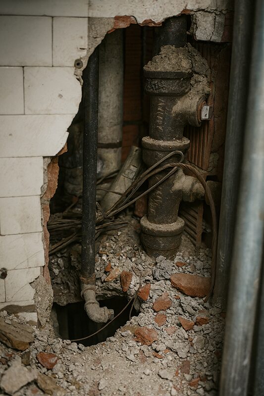Exposed pipes and damaged wall with debris during a building inspection, highlighting potential asbestos and hazardous material risks