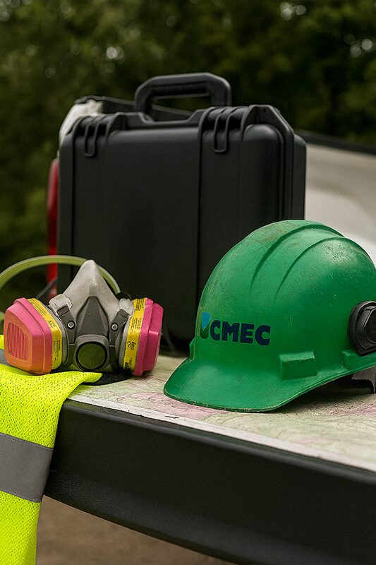 CMEC-branded green hard hat, respirator mask, and high-visibility safety vest placed on a work surface next to a black equipment case.