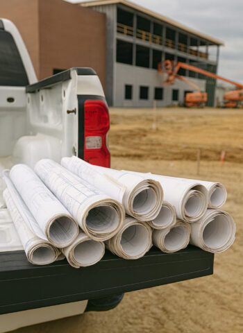Rolled construction blueprints stacked on the tailgate of a white pickup truck at a building site with a partially constructed structure in the background